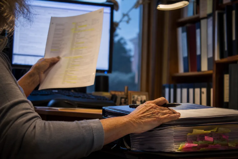 A hand holding a printed VA accreditation record page next to a monitor while a claims agent checks a representative line by line